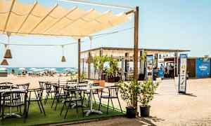 En strandbar med uteservering under et seiltak, med utsikt over stranden og havet. (AI-generert tekst)