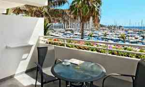 Utsikt fra en balkong med et glassbord og to stoler som vender mot en marina og havet. (AI-generert tekst)