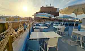 Bildet viser en takterrasse med bord, blå og hvite stoler samt parasoller under en solrik himmel. (AI-generert tekst)