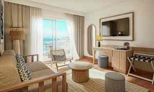 Et lyst og moderne hotellrom med utsikt over havet. Det er en sofa, et sofabord, en stol og en TV på veggen. (AI-generert tekst)
