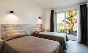 To enkeltsenger i et lyst rom med store vinduer mot en balkong og utsikt over palmer. (AI-generert tekst)