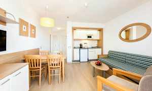 Et moderne hotellrom med en liten kjøkkenkrok, en spiseplass med bord og stoler samt en sittegruppe med sofa og salongbord. (AI-generert tekst)