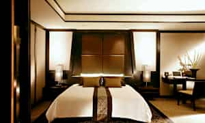 Et moderne soverom med en stor seng i midten, omgitt av elegante, mørke trepaneler og varme lyskilder. (AI-generert tekst)