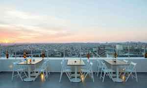 En takterrasse med bord og stoler og utsikt over en by ved solnedgang. (AI-generert tekst)