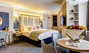 Lyst hotellrom med dobbeltseng, lenestoler og fruktskål. (AI-generert tekst)