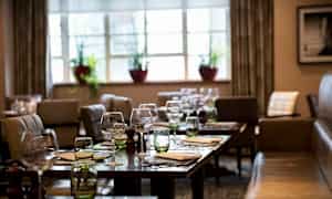 Et restaurantområde med oppdekkede bord, glass og servietter. I bakgrunnen er vinduer med planter. (AI-generert tekst)