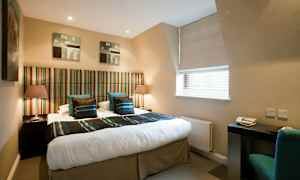 Et hotellrom med en dobbeltseng med stripete sengegavl og to nattbord med lamper. Et skrivebord med stol ved et vindu. (AI-generert tekst)