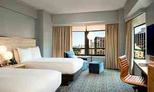 Et hotellrom med to dobbeltsenger, store vinduer med utsikt over byen, en lenestol og et skrivebord med en stol. (AI-generert tekst)