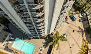 En høy bygning sett ovenfra med balkonger som vender utover. Nedenfor er det et basseng, solsenger og parkerte biler. (AI-generert tekst)