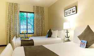 Et hotellrom med to brede senger, hver med en pute og et teppe. Et vindu med gardiner og utsikt over trær. (AI-generert tekst)