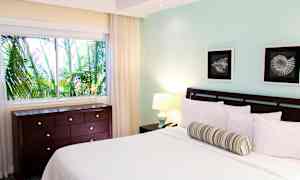 Et soverom med en seng, to malerier, et vindu med utsikt mot frodige palmer og en kommode. (AI-generert tekst)