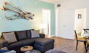 En moderne stue med en grå hjørnesofa, et glassbord og en sitteplass med mønstrede puter. En dekorativ metallskulptur henger på en lyseblå vegg. (AI-generert tekst)