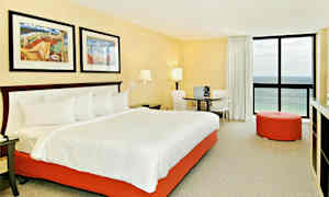 Et hotellrom med en stor seng som har hvitt sengetøy og en oransje sengebunn. Rommet har to malerier over sengen, et rundt bord med to stoler, en oransje sittepuff og et vindu med havutsikt. (AI-generert tekst)