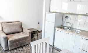 Et kjøkken og stue med en beige lenestol, et sofabord, et kjøleskap og en oppvaskkum. (AI-generert tekst)