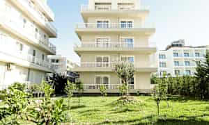 Et flerfamiliehus i lys farge med en hage med plen og små trær i forgrunnen. (AI-generert tekst)