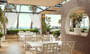 Utendørsområde med hvite bord og stoler under et tak, omgitt av grønn vegetasjon og havutsikt. (AI-generert tekst)