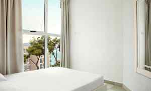 Et soverom med en hvit seng nær et stort vindu med utsikt over sjøen og nærliggende bygninger. (AI-generert tekst)