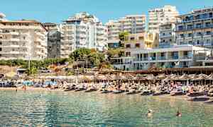 En strand med solsenger og parasoller foran flere moderne bygninger. (AI-generert tekst)