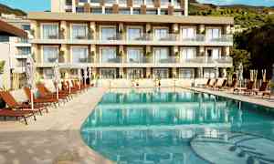 Bilde av et svømmebasseng med solsenger og parasoller foran et moderne hotell med balkonger. (AI-generert tekst)