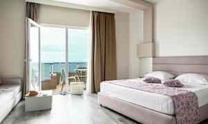 Et hotellrom med en stor seng, glassdører ut til en balkong og utsikt over havet. (AI-generert tekst)