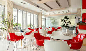 En moderne kafé med røde og hvite stoler og bord, plassert i et lyst rom med store vinduer og innendørs planter. (AI-generert tekst)