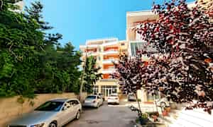 Bildet viser en boligblokk med parkerte biler foran, omgitt av busker og trær. (AI-generert tekst)