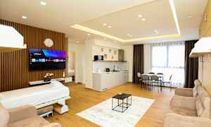 Et moderne hotellrom med åpen planløsning som inkluderer en sovesofa, TV, kjøkkenområde, spisebord og store vinduer med gardiner. (AI-generert tekst)