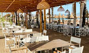 Bilde av en utendørs servering med trebord og hvite stoler under en trepergola. (AI-generert tekst)