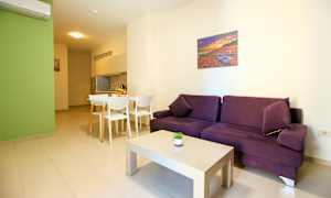 En lyst møblert stue med en lilla sofa, hvit sofabord og spisekrok ved siden av et kjøkken. (AI-generert tekst)