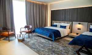 Et moderne hotellrom med to senger, en sittegruppe med to stoler og et bord, samt mørke gardiner ved vinduet. (AI-generert tekst)