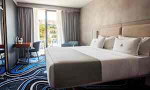 Et hotellrom med en oppredd dobbeltseng, et skrivebord med stol og vindu mot balkong. (AI-generert tekst)