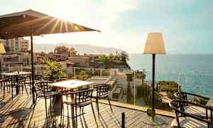 Terrasse med spisebord og stoler, havutsikt og bygninger i bakgrunnen. (AI-generert tekst)