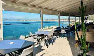 Utendørs servering med bord og stoler nær sjøen, med utsikt over vannet og en by i bakgrunnen. (AI-generert tekst)