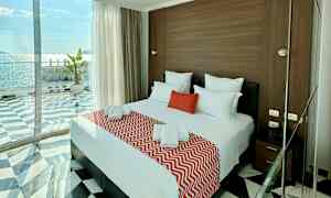 Et hotellrom med en dobbeltseng dekket med røde og hvite tekstiler, store glassvinduer med havutsikt. (AI-generert tekst)