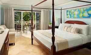 Rommet har en seng med fire stolper og er innredet med lyse møbler. En balkong med utsikt over frodige palmer er synlig gjennom de åpne glassdørene. (AI-generert tekst)