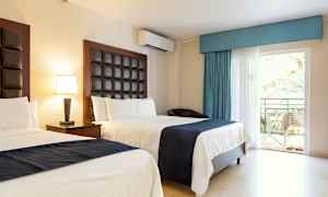 Hotellrom med to senger, lyse vegger og mørke sengegavler. Bord med lampe, og en balkongdør med utsikt over grøntområde. (AI-generert tekst)