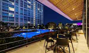 Utsikt over et opplyst basseng fra en terrasse med bord og stoler, omgitt av vegetasjon og med en bygning i bakgrunnen om kvelden. (AI-generert tekst)