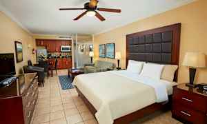 Et hotellrom med en stor seng, nattbord, polstret lenestol, spiseplass og et kjøkken i bakgrunnen. (AI-generert tekst)