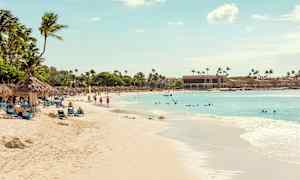 En sandstrand med palmer og solsenger, folk bader og går tur, og havet strekker seg ut under en klar himmel. (AI-generert tekst)