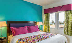 Et soverom med en dobbeltseng, fargerikt sengetøy og puter. Veggene er turkise, og rommet har vinduer med utsikt over palmer og havet. (AI-generert tekst)