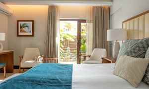 Et lyst og moderne soverom med en seng oppredd med hvitt og blått sengetøy, to stoler og et lite bord ved en glassdør med utsikt over en hage. (AI-generert tekst)