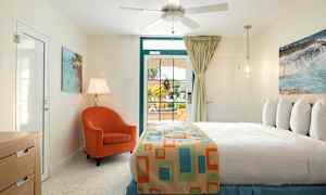 Et soverom med lys innredning. En seng med et fargerikt sengeteppe, en oransje lenestol, bordlampe og et vindu med utsikt ut. (AI-generert tekst)