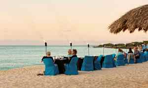 Strand med dekket bord og sitteplasser med blå stoltrekk, hvor folk spiser og sosialiserer. Havet og himmelen kan ses i bakgrunnen. (AI-generert tekst)