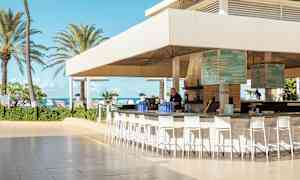 En utendørs bar med tak og høye stoler, som ligger nær stranden med palmer og blå himmel i bakgrunnen. (AI-generert tekst)