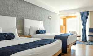 Et hotellrom med to senger med hvite laken og blå sengetepper. Vinduet er dekket med blå gardiner og lys strømmer inn i rommet. (AI-generert tekst)