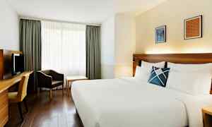 Et hotellrom med en stor seng med hvitt sengetøy, et skrivebord med stol, en lenestol og et bord ved et vindu med gardiner. (AI-generert tekst)