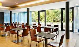 En lys spisestue med bord og stoler ved store vinduer med utsikt over grønne busker. (AI-generert tekst)