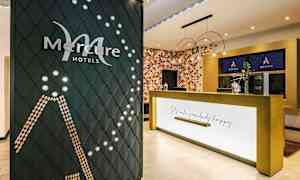 Resepsjonen på et Mercure-hotell med selskapets logo synlig på en dekorativ vegg og en resepsjon med teksten "Let's make somebody happy". (AI-generert tekst)