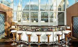 En cocktailbar med høye stoler foran en glassvegg med utsikt over en bygning med fargerike takdekorasjoner. (AI-generert tekst)