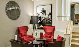 To røde lenestoler står rundt et rundt bord, ved siden av en gulvlampe og foran en vegg dekorert med kunst i et hotellinteriør. (AI-generert tekst)